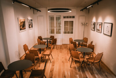 Modern eingerichteter Raum eines Cafés mit Holzboden, runden Tischen und Holztstühlen. An den Wänden hängen Kunstwerke, die in warmem Licht beleuchtet sind.